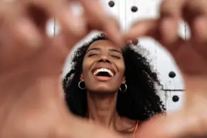 Optimistic young African American female laughing happily and showing heart shaped sign while standing against old wall