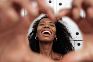 Optimistic young African American female laughing happily and showing heart shaped sign while standing against old wall