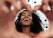 Optimistic young African American female laughing happily and showing heart shaped sign while standing against old wall